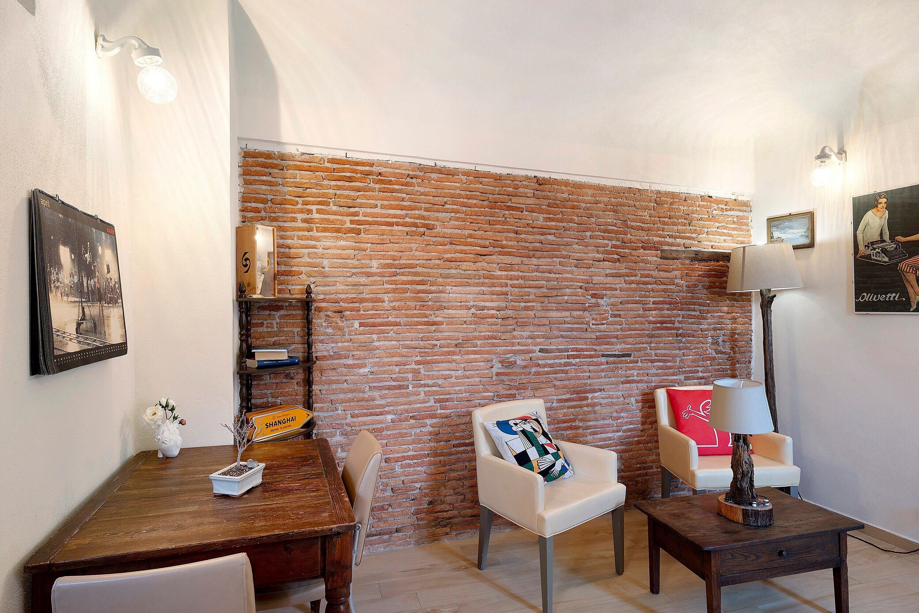 Ein Zimmer mit Backsteinwand, einem Holztisch, einem weißen Sessel, einem kleinen Tisch mit Lampe und einem Bücherregal.