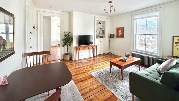 A living room with a wooden floor, a green sofa, a coffee table, a TV, a bookshelf, and a window with a view. - Downtown Brunswick 1st FL Sun Drenched With Porch (Brunswick)