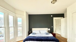 A bedroom with a large bed, a nightstand, a lamp, and a view through the window. - Downtown Brunswick 1st FL Sun Drenched With Porch (Brunswick)