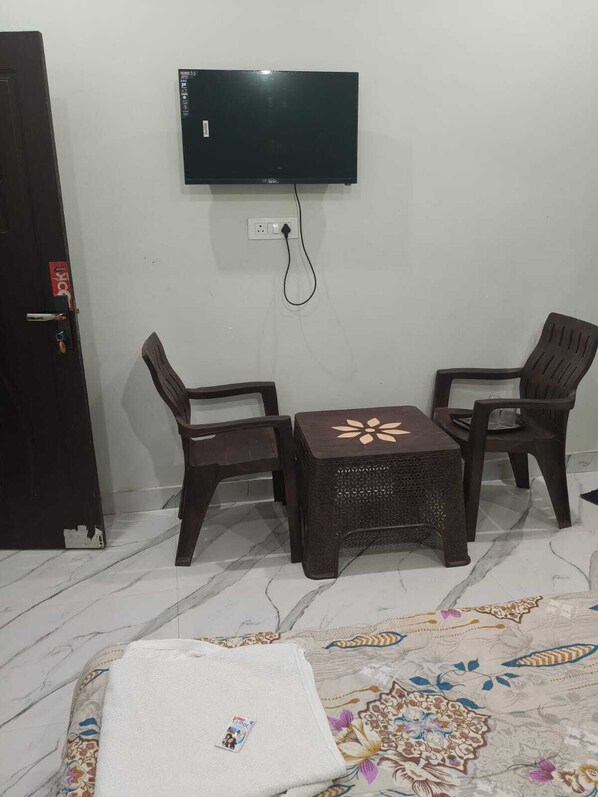 Une pièce avec un téléviseur fixé au mur, deux chaises en bois, une petite table, une porte avec une serrure et une couvre-lit à motifs.