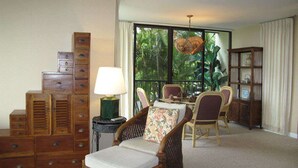 Quarto com cadeira de vime, penteadeira de madeira, mesa com cadeiras e uma grande janela com vista para o verde.