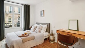 Een slaapkamer met een bed, een bureau, een stoel, een raam met gordijnen en uitzicht op gebouwen.