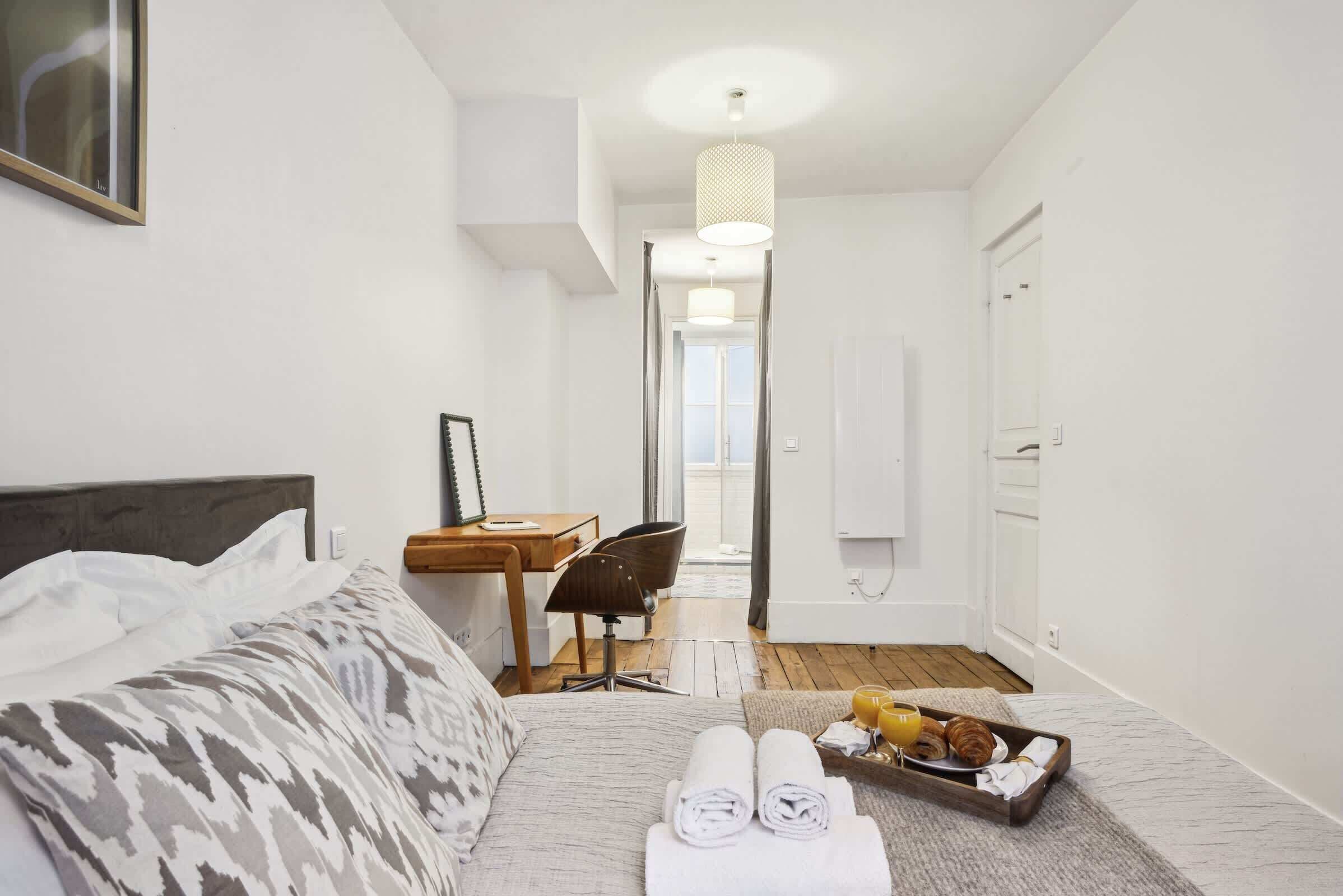 Een hotelkamer met een bed, een bureau, een stoel, een dienblad met handdoeken en croissants, en uitzicht op het balkon.