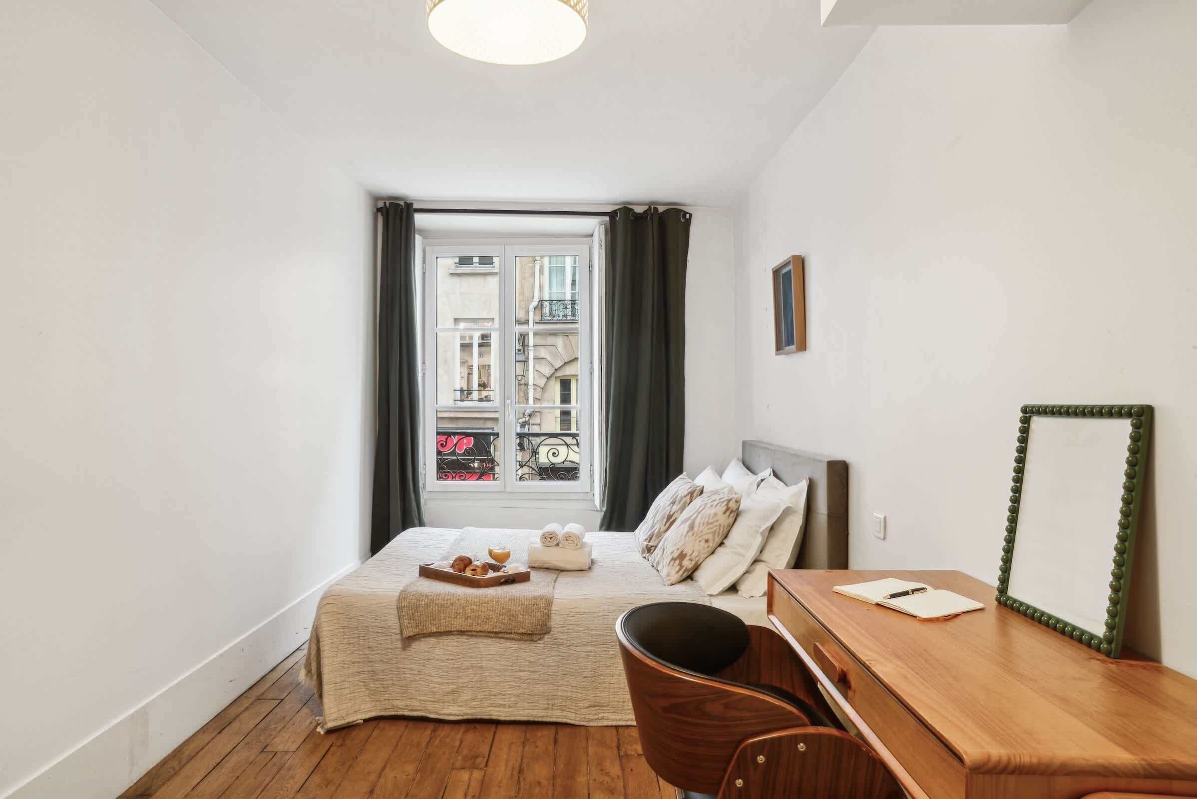 Una camera da letto con un letto, una scrivania in legno, una sedia, una finestra con tende e vista sugli edifici esterni.