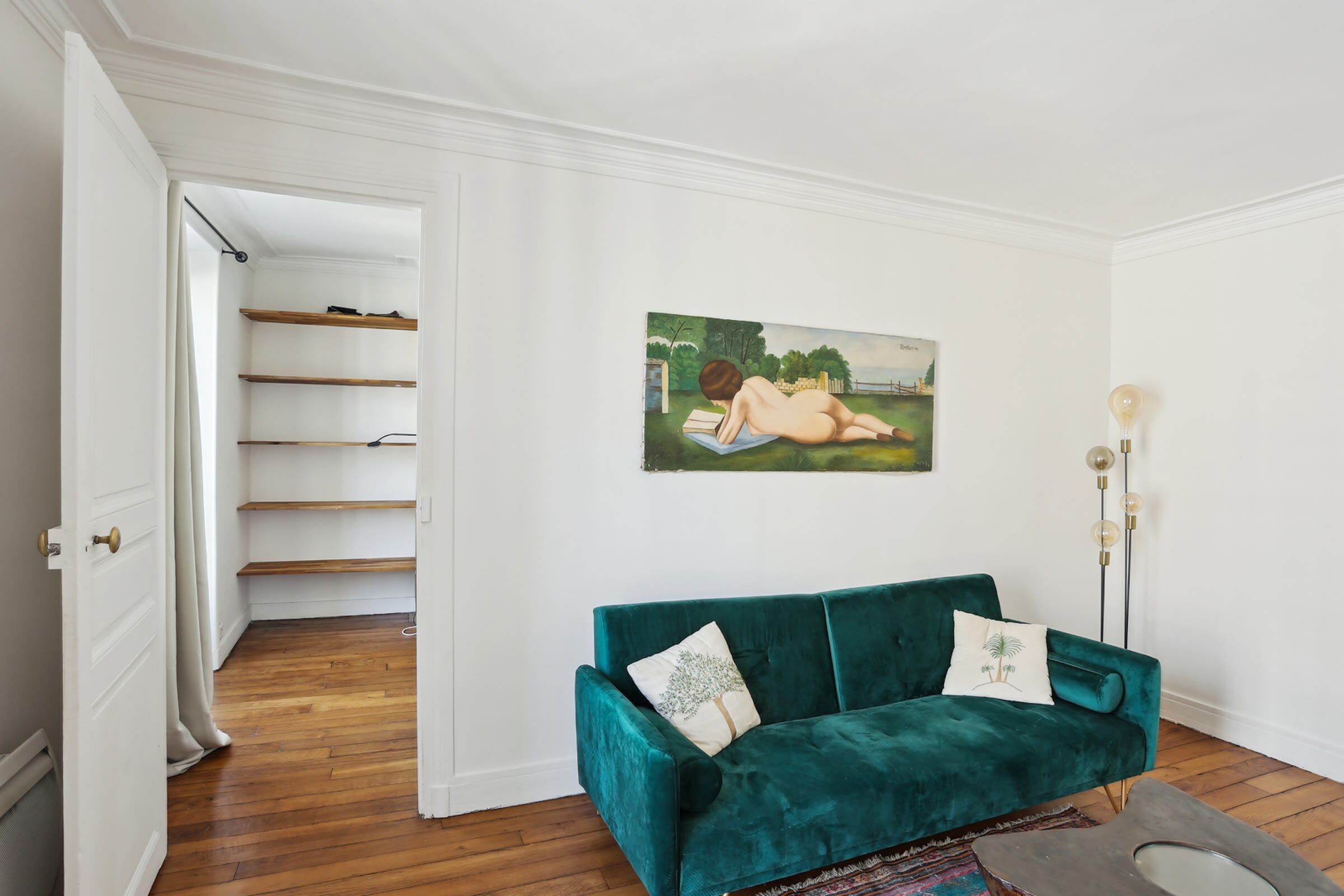 A room with a teal velvet sofa, a painting of a nude figure, and a wooden floor.