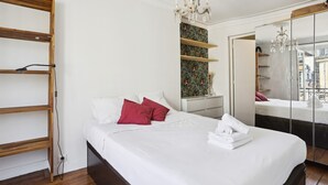 Una cama bien hecha con almohadas blancas y rojas, una estantería de madera, un espejo decorativo y una araña.