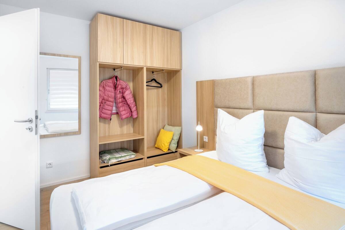A hotel room with a bed, a wooden wardrobe, a bedside table with a lamp, and a window with blinds.
