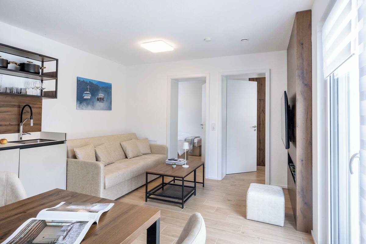 A modern living room with a sofa, coffee table, and a dining area with a table and chairs. There is a kitchen area with a sink and cabinets, and a TV mounted on the wall.