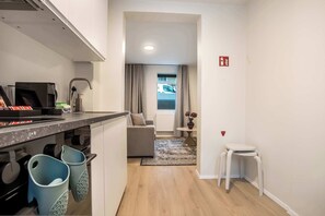 A modern kitchen with a countertop, sink, and cabinets. There is a dining area with a sofa and a small table. A hallway leads to a living room with a view of a parked car outside. - TYS One Bedroom Basement Apartment (Reykjavík)
