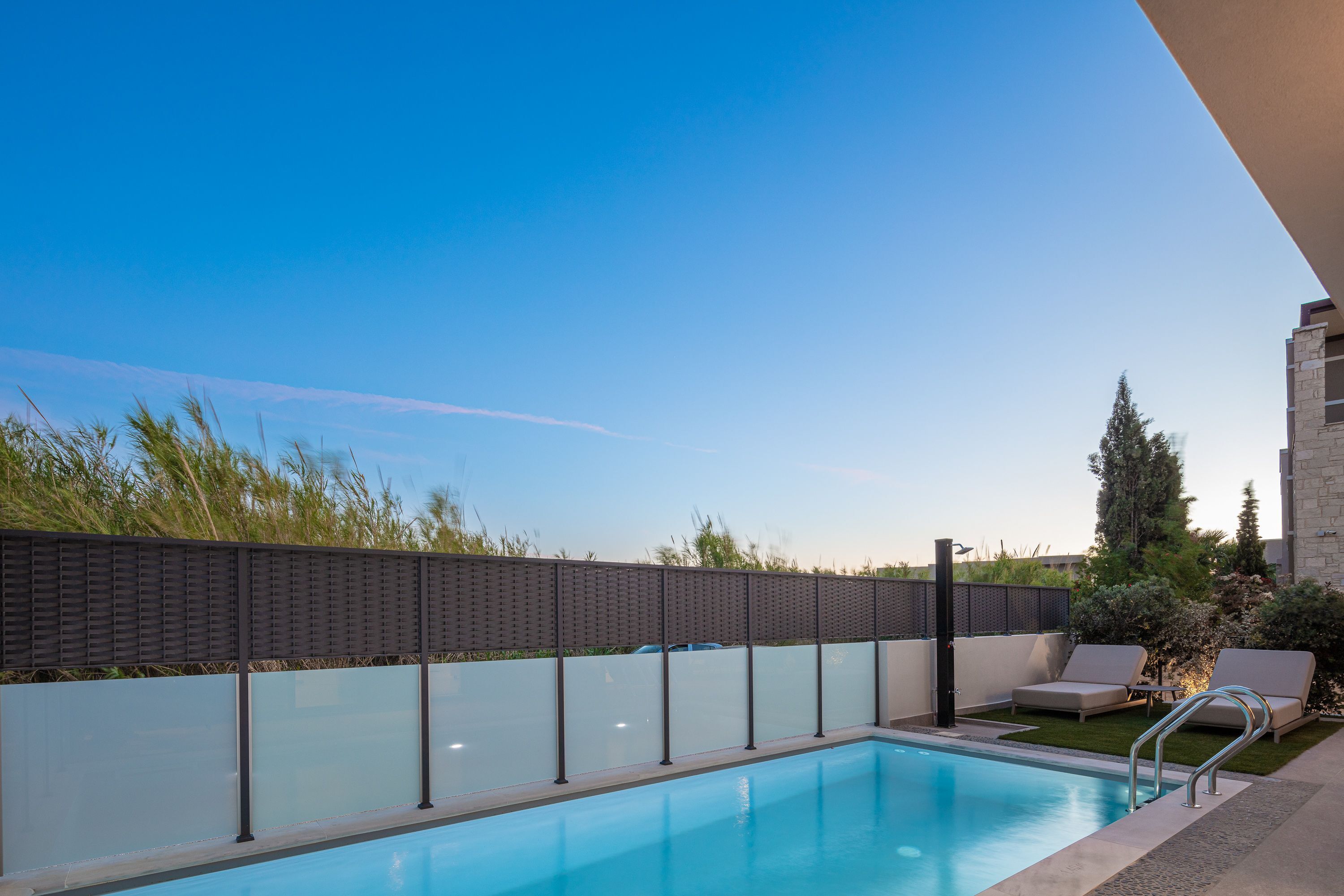 Una piscina con pared de cristal, sillas de descanso y un edificio de piedra al fondo.
