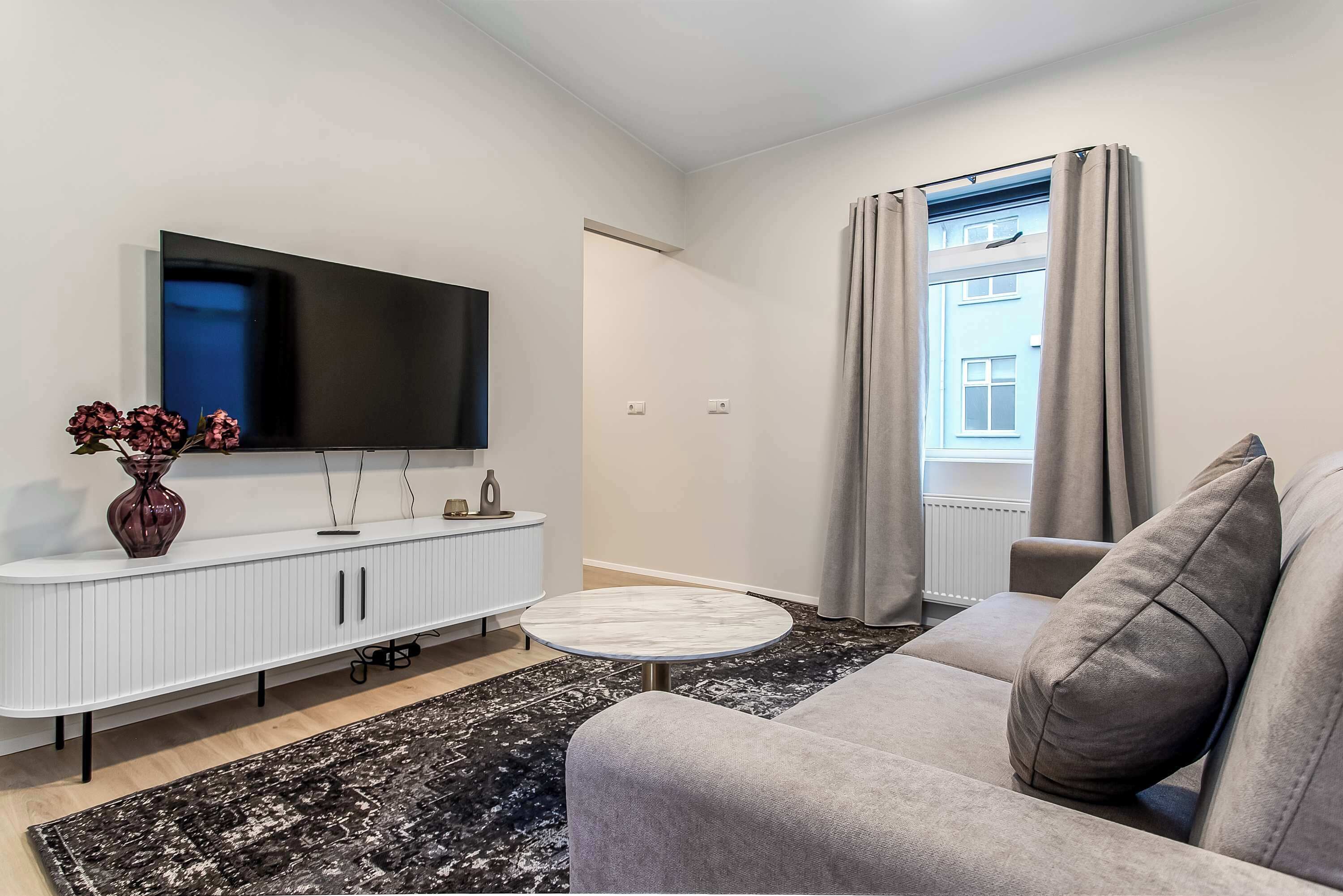 A modern living room with a flat-screen TV, a white console table, a grey sofa, a round coffee table, and a black and white patterned rug.