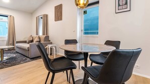 A modern living room with a sofa, a dining table with chairs, a large window with curtains, and a framed picture on the wall. - TYS One Bedroom Apartments (Reykjavík)