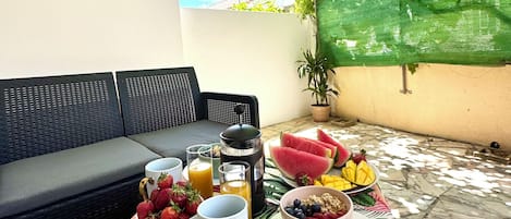 Una mesa de comedor con alimentos para el desayuno, un sofá negro y un árbol que da sombra.