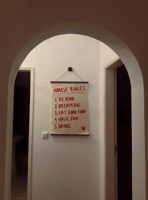 Een gang met een bord waarop 'Huisregels' staan: '1. Wees aardig', '2. Droom groot', '3. Eet lekker eten', '4. Veel plezier', en '5. Dans'.