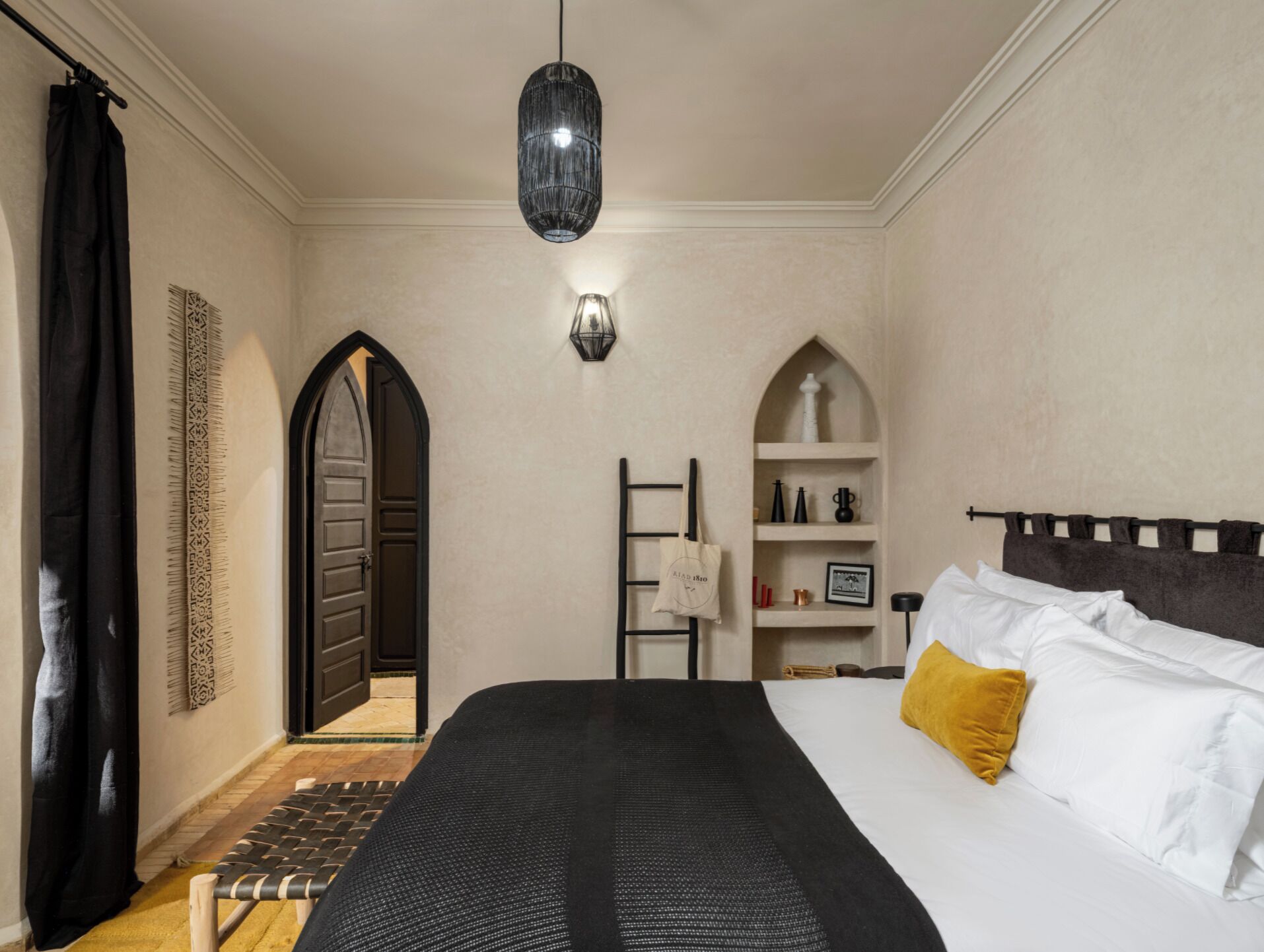 Une chambre à coucher avec un lit, un luminaire suspendu, une porte, une étagère avec des objets et un tapis à motifs.