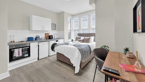 A compact living space with a kitchenette, a bed, a dining area, and a window with blinds. - Sunny Serenity: Studio Living with a Breeze (London)
