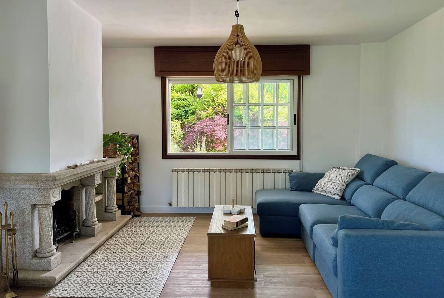 En stue med pejs, en blå sofa, et sofabord og et vindue med udsigt til grønne omgivelser.