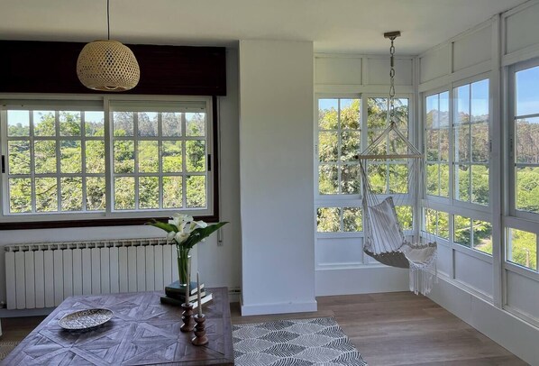 A room with large windows, a hanging chair, a table with a vase of flowers, and a textured rug. - Casa Santi - 10 Min de Santiago  (Santiago de Compostela)