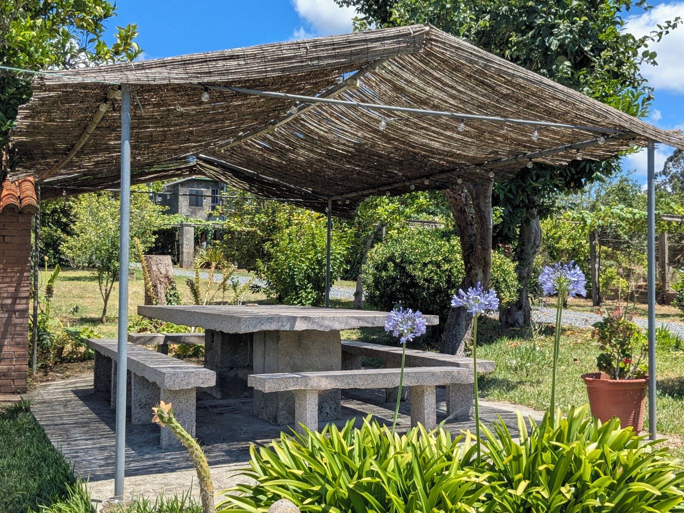 A covered outdoor seating area with a table and benches, surrounded by greenery and trees.