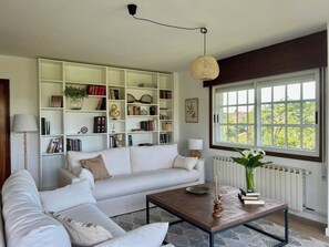 A living room with a white sofa, a coffee table, a bookshelf, and a window with a view. - Casa Santi - 10 Min de Santiago  (Santiago de Compostela)