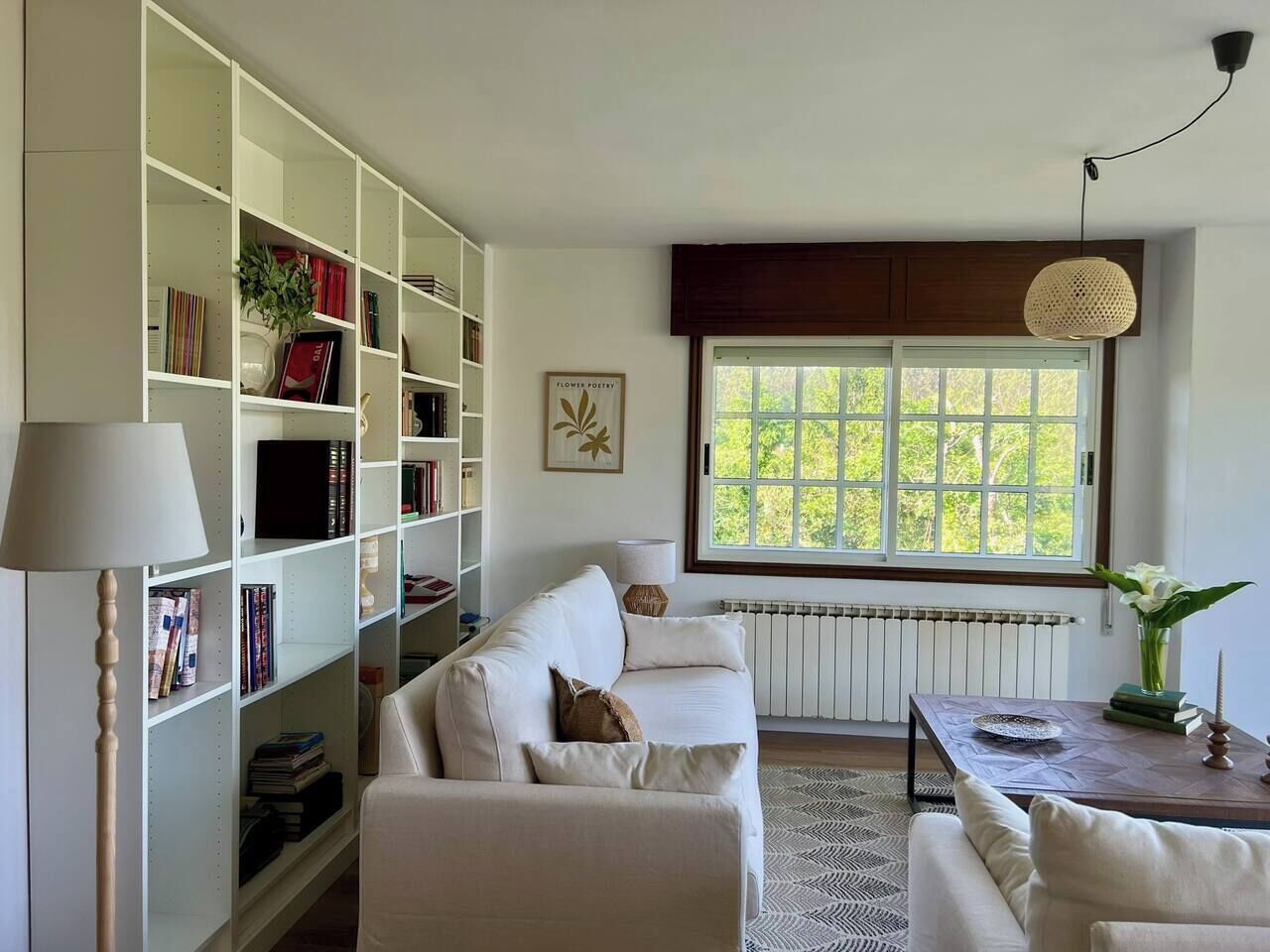 A living room with a white sofa, a bookshelf, a table with a vase, and a window with a view of greenery.