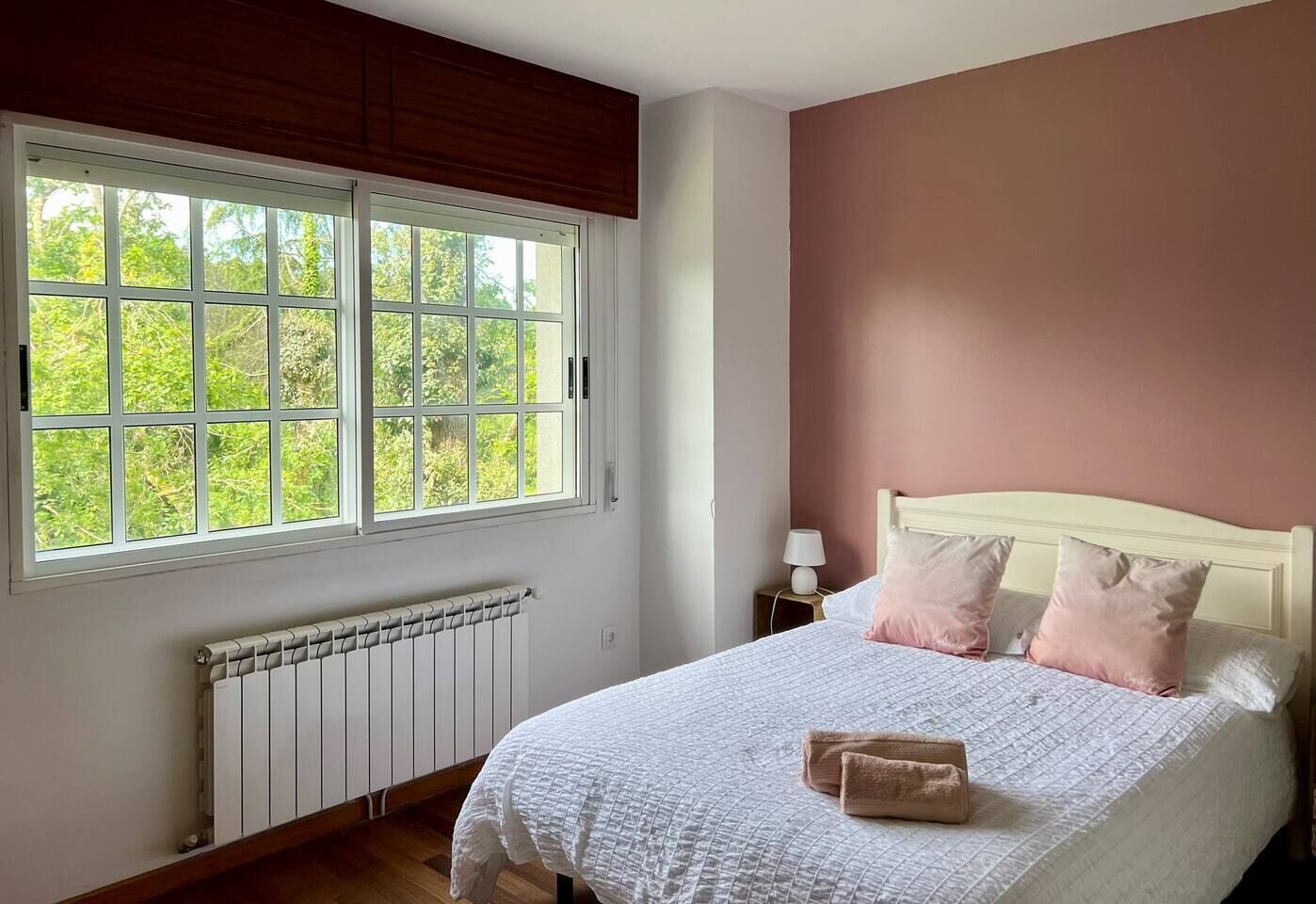 A bedroom with a large window, a radiator, a bed with pillows, and a bedside table with a lamp.