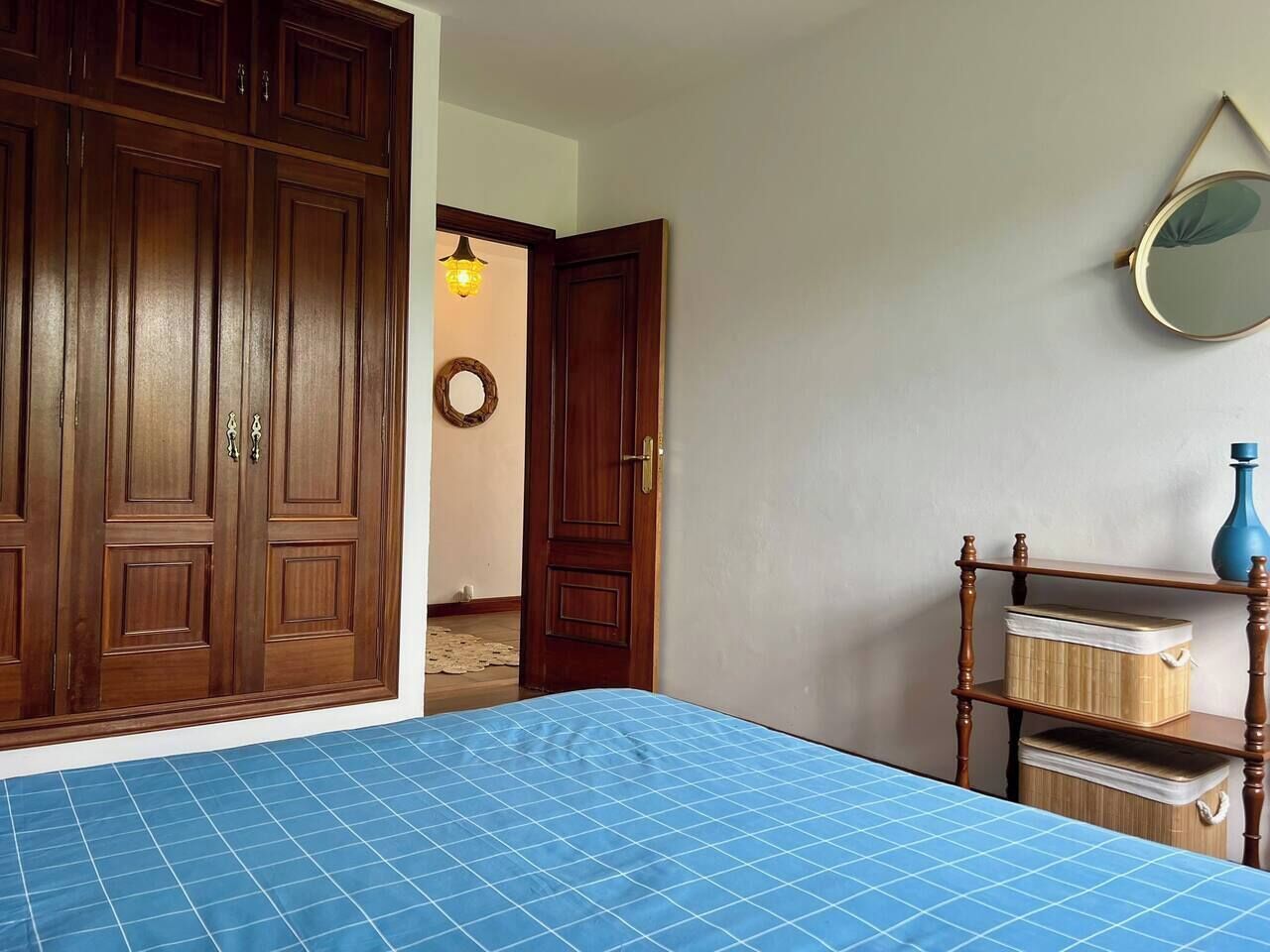Una stanza con un armadio capiente, un letto con copriletto a quadretti blu, uno scaffale in legno con contenitori e uno specchio appeso al muro.