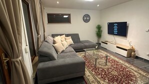Un soggiorno moderno con un divano angolare grigio, una TV a schermo piatto montata a parete, un tappeto decorato e una piccola pianta in vaso.