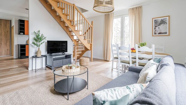 A modern living room with a wooden staircase, a round glass coffee table, a grey sofa, and a dining area with white chairs. - Imperial Apartments - Andersa (Sopot)