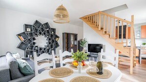 A modern living room with a dining table, a geometric wall art piece, a staircase, and a TV in the background. - Imperial Apartments - Andersa (Sopot)