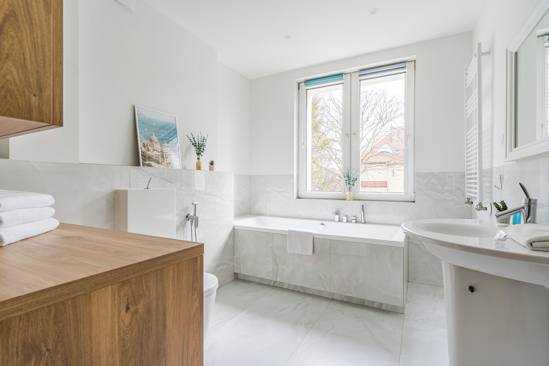 Un baño moderno con bañera, lavamanos e inodoro. Se ve un ventanal con vistas a árboles y edificios.