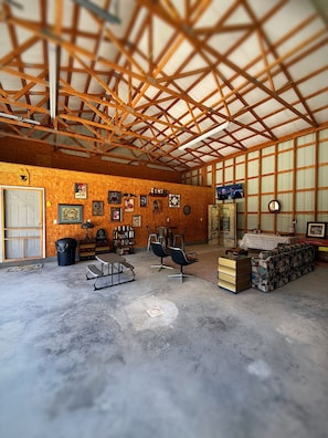 A spacious barn interior with a high ceiling, wooden beams, a concrete floor, and various items including a table, chairs, a sofa, and a television.