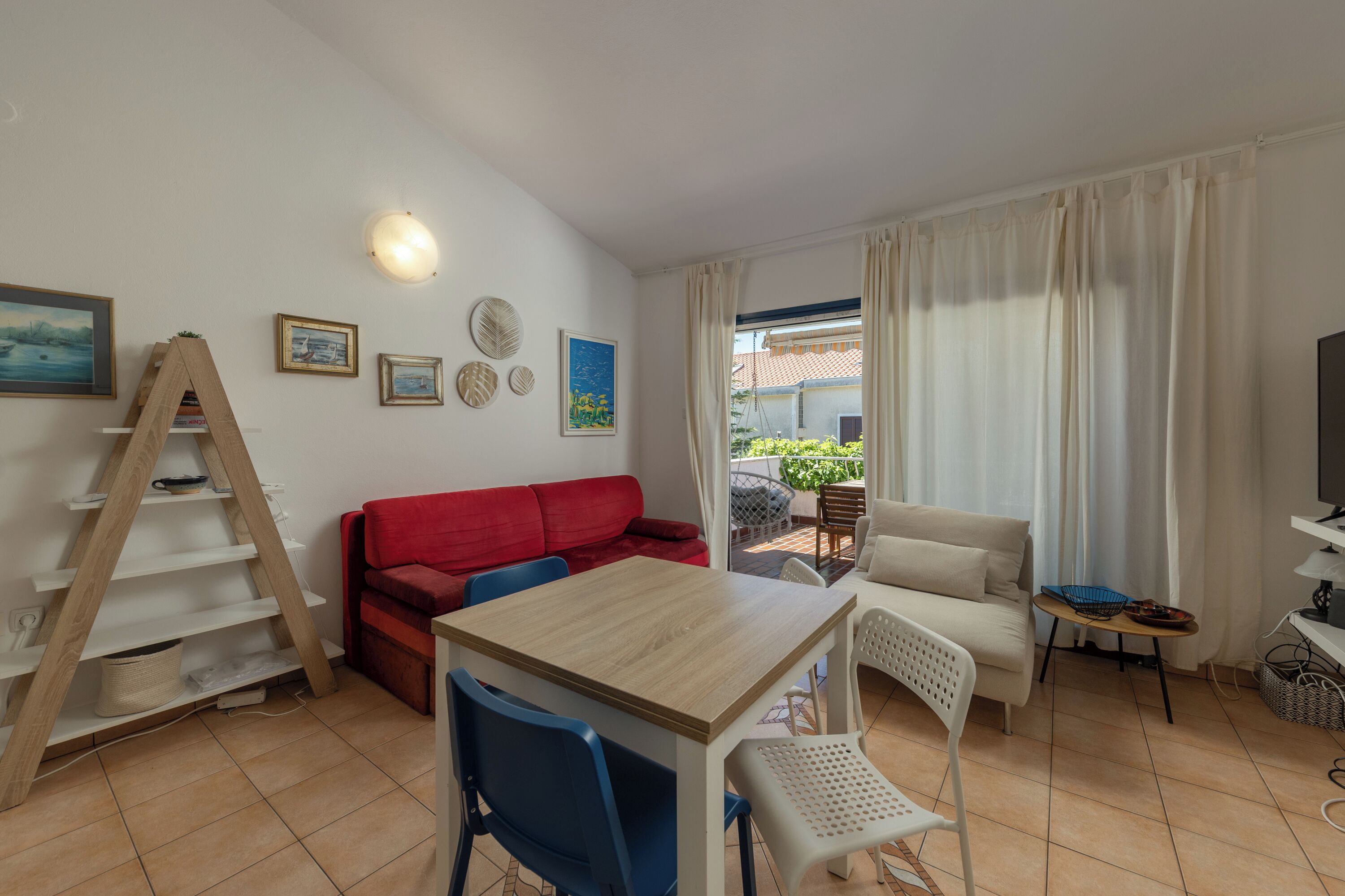 A furnished living room with a wooden table, two sofas, a TV, and a balcony with plants.