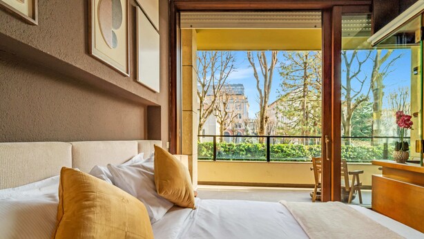 Habitación de hotel con una cama grande, un balcón con vistas a edificios y árboles, y un escritorio de madera con un florero.