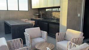 Ett modernt utomhusområde med sittplatser, bestående av flätade stolar och ett runt bord, ett kök med ett stort fönster och utsikt över stadens silhuett.