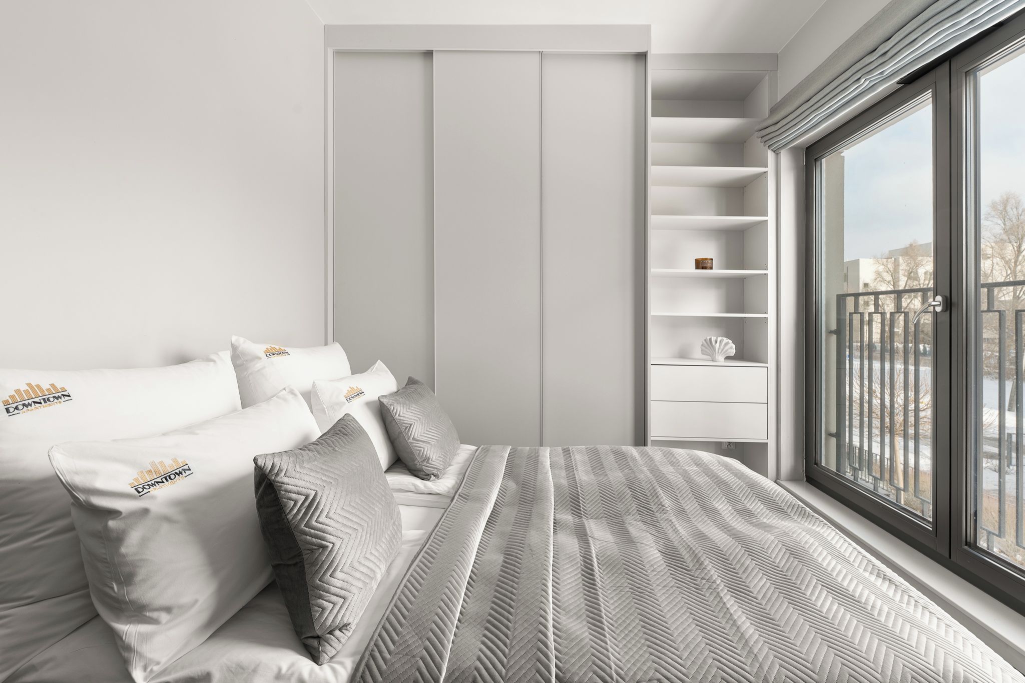 Ein modernes Schlafzimmer mit einem Bett, weißen Kissen, einer grau gemusterten Tagesdecke, einem Einbauschrank und einem Fenster mit Ausblick.