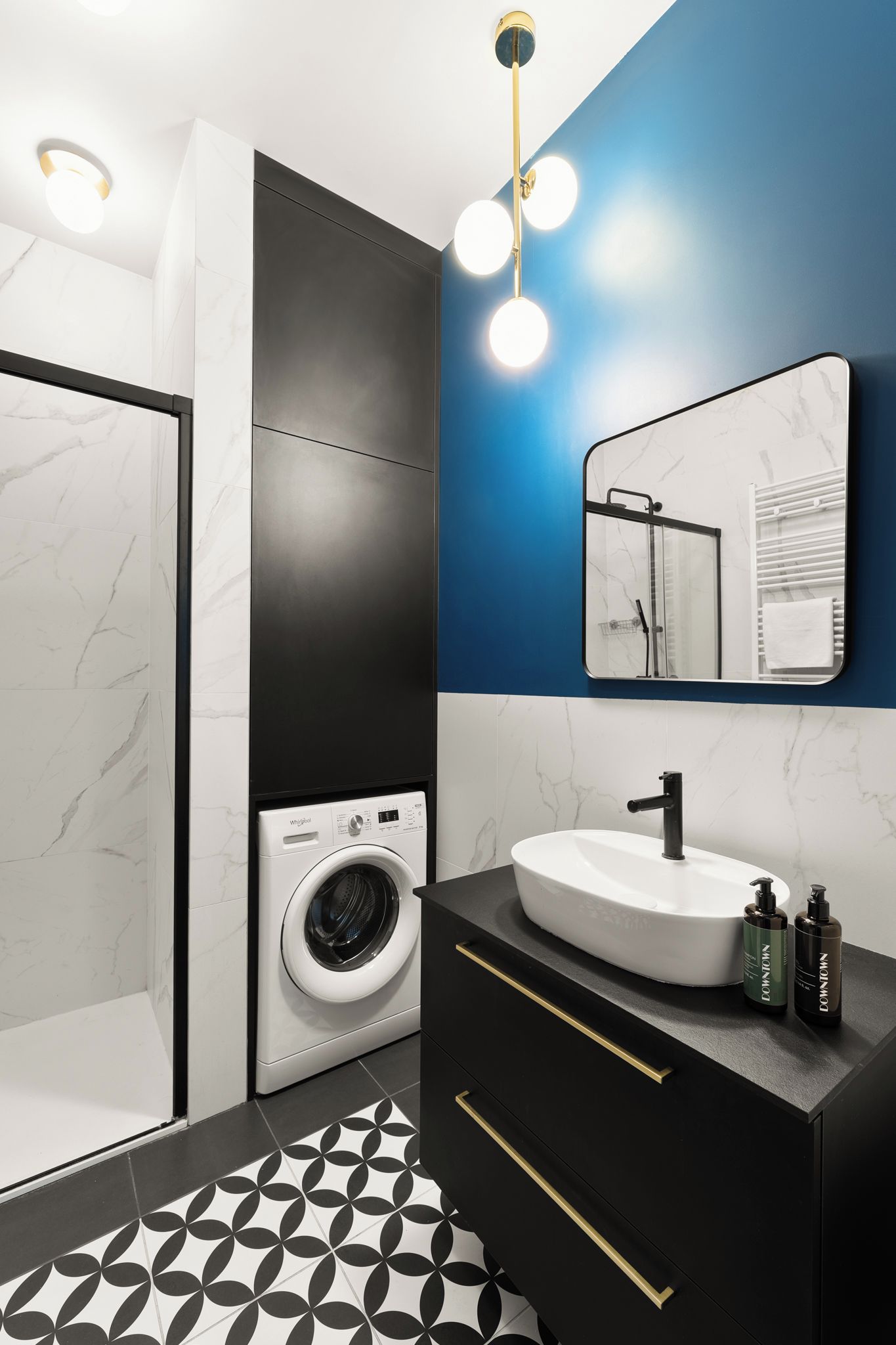 Un baño moderno con suelo de mármol, lavadora, lavabo con espejo y mueble de tocador negro con tiradores dorados.
