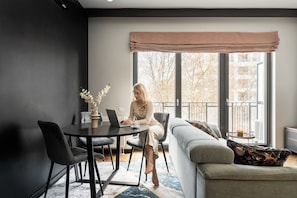 A modern living room with a dining area, large windows with a view of trees, and a person working on a laptop. - Art Deco Mokotów Suites - Parking & Gym by Downtown Apartments (Warszawa)