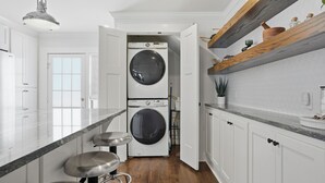 A modern kitchen with a built-in washer and dryer, bar stools, white cabinetry, and a wooden shelf. - Midtown 3-bedroom house in vibrant Tulsa close to TU, Downtown, EXPO Square (Tulsa)