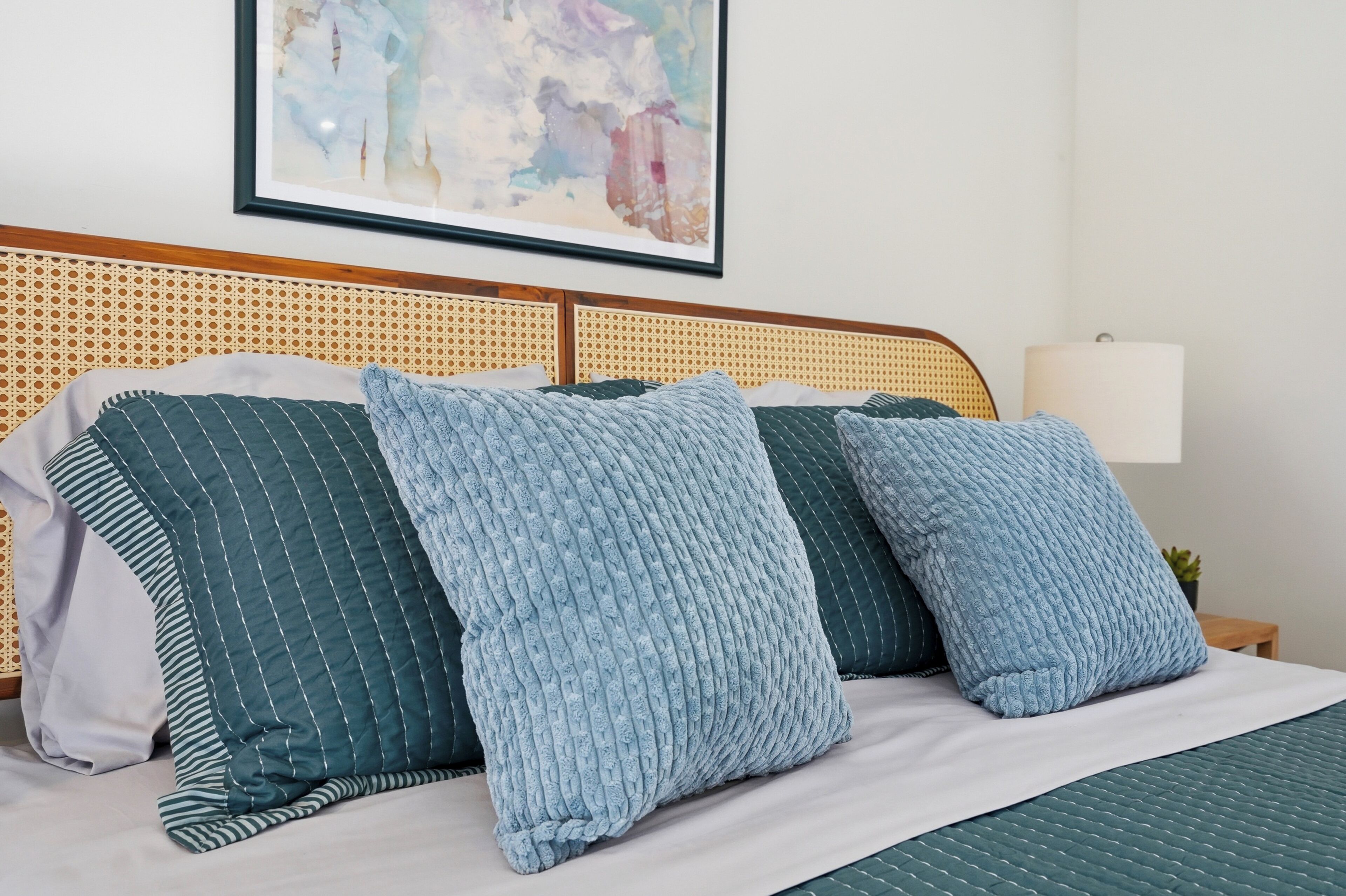 A bed with striped and textured pillows, a woven headboard, a framed abstract painting, and a bedside lamp.