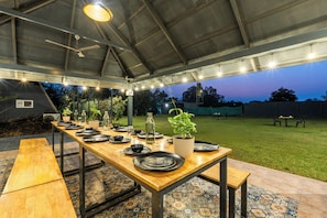 An outdoor dining area with a long wooden table set for a meal, surrounded by chairs, under a covered structure with a ceiling fan and string lights. - Alaya Stays Inara Retreat Cottage (Bengaluru)