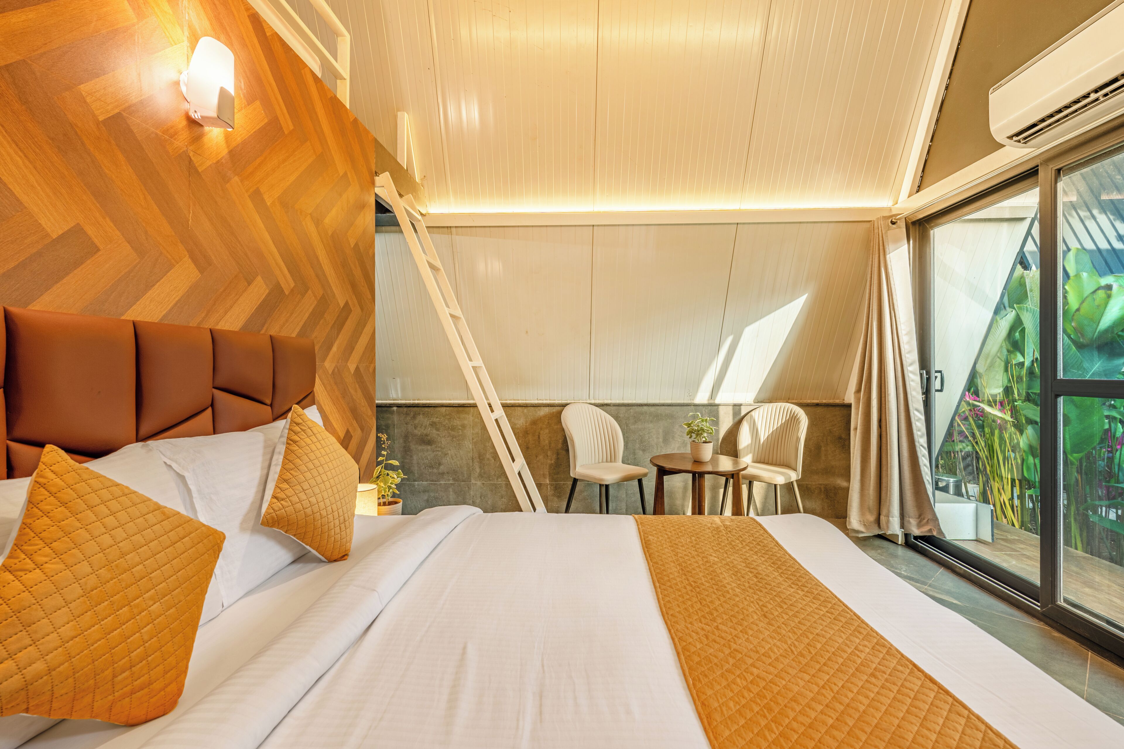 A modern hotel room with a large bed, a wooden headboard, a small table with chairs, and a view of greenery through the window.