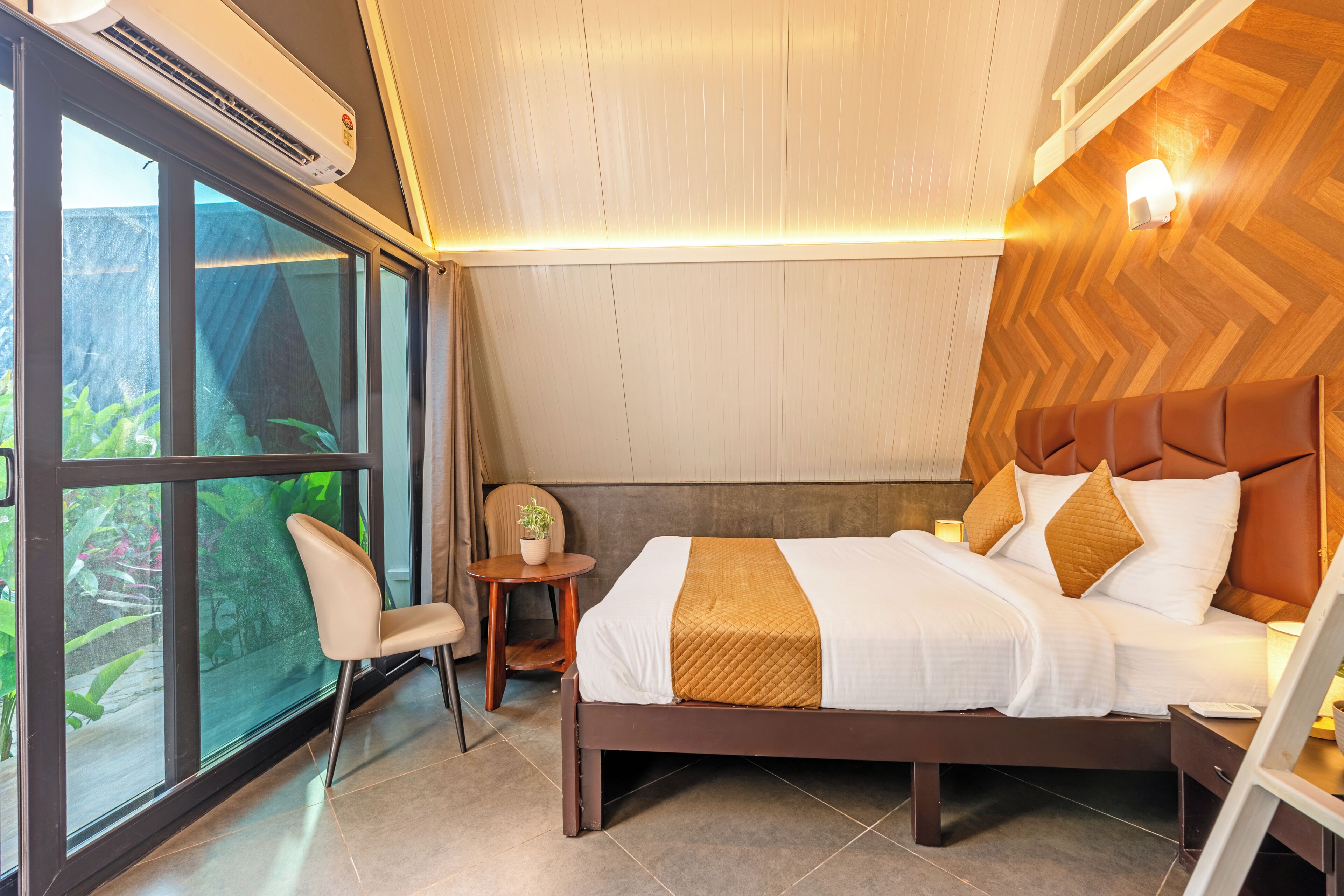 A hotel room with a bed, a chair, a small table, and a window with a view of greenery.