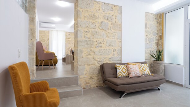 Un espace de vie avec un mur en pierre, un canapé, un fauteuil et un escalier menant à une autre pièce.