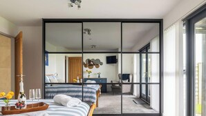 A modern hotel room with a large sliding glass door, a bed with striped bedding, a bedside table with a vase of flowers, and a view of the adjacent room.