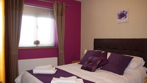 A bedroom with a bed, purple bedding, a headboard, a window with curtains, and a wall art piece.