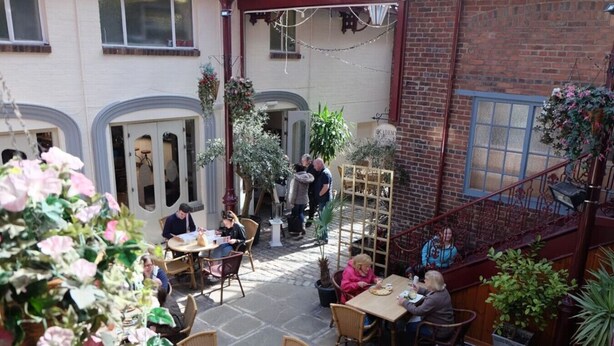 An outdoor café with tables and chairs, surrounded by potted plants and a brick wall.