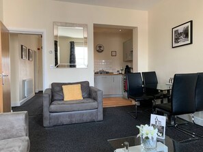 Un salon avec un canapé gris, une table basse et un miroir. Au fond, on aperçoit un coin cuisine avec une table à manger et des chaises.