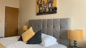 Une chambre d’hôtel avec une tête de lit capitonnée, un lit recouvert d’une literie blanche et grise, deux lampes dotées d’abat-jour à motifs et un tableau représentant des ânes accroché au mur.