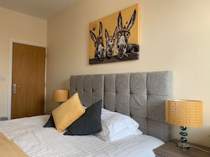 Une chambre d’hôtel avec une tête de lit capitonnée, un lit recouvert d’une literie blanche et grise, deux lampes dotées d’abat-jour à motifs et un tableau représentant des ânes accroché au mur.